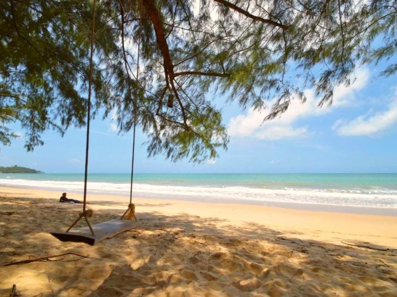 Een verlaten strand op Koh Lanta met zacht zand en helderblauw water. Op de voorgrond hangt een houten schommel aan een boomtak en werpt schaduw op het zand. In de verte zit een persoon in alle rust naar de zee te kijken. Een rustige, idyllische plek onder de schaduw van groene naaldbomen.