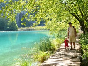 Man en peuter wandelen over een houten vlonderpad langs een turquoise meer omringd door groene bomen in Kroatië