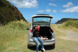 Lachende vrouw geeft een duim omhoog vanuit de open kofferbak van een zilveren auto tijdens een picknick met een kustlandschap op de achtergrond in Nieuw-Zeeland