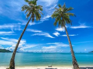 Strand met twee hoge palmbomen met lampjes aan de stammen, een schommel tussen de bomen, een paar sluierwolken in de lucht en eilanden in de verte