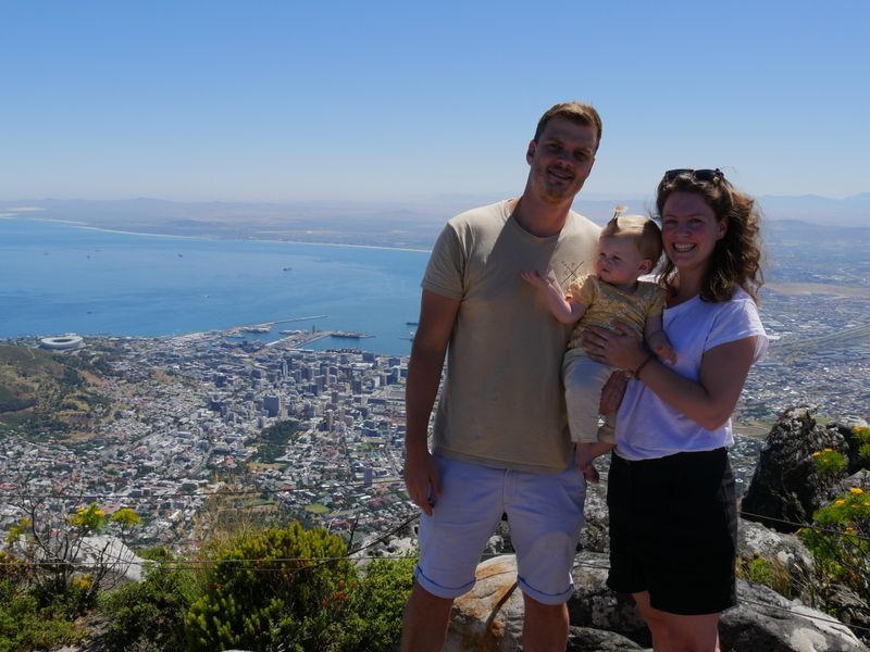 Gezin van drie met man, vrouw en baby lacht op een bergtop met uitzicht over de stad en oceaan in Zuid-Afrika