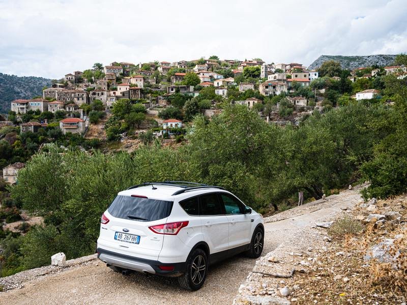 Witte SUV op een onverharde weg aan de Albanese Rivièra met een heuveldorp van traditionele huizen op de achtergrond