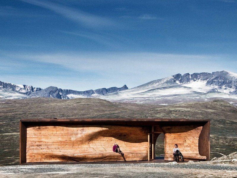 Two people sit inside a curved wooden shelter overlooking a vast, snow-capped mountain landscape.