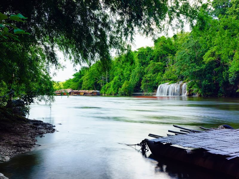 Een rustige rivier slingert zich door het groen, met aan de overkant een kleine waterval en traditionele drijvende huisjes. Op de voorgrond zie je een bamboevlot aan de oever.