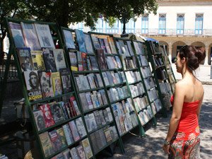 Vrouw in een rode top bekijkt een boekenkraam buiten met veel boeken, waaronder titels met Che Guevara, in Cuba