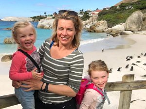 Vrouw met twee lachende kinderen op een zandstrand met blauw water, rotsen en een kolonie pinguïns in Zuid-Afrika
