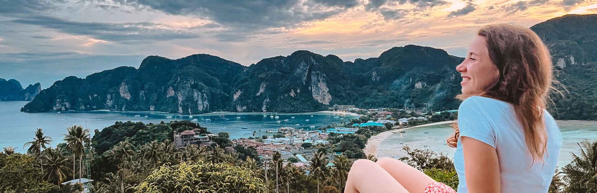 Een reizigster zit op een rotspunt en kijkt uit over de baai van Koh Phi Phi, met steile kalksteenrotsen, palmbomen en een dorpje aan zee. De lucht kleurt zachtroze en oranje. Ze draagt een wit shirt en roze short, en glimlacht ontspannen.