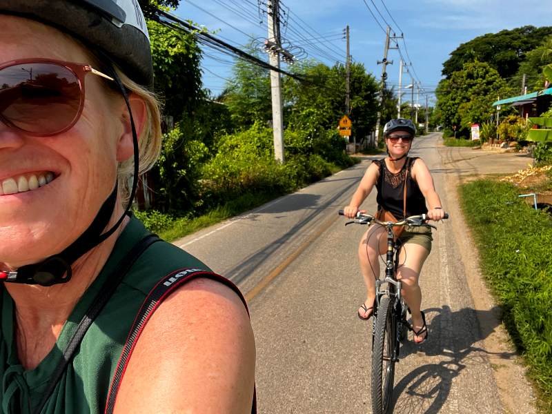 Twee vrouwen fietsen over een rustige weg met helmen op, omringd door groen en elektriciteitspalen.