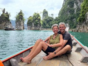 Twee reizigers zitten samen op de punt van een longtailboot op het water tussen kalksteenrotsen.
