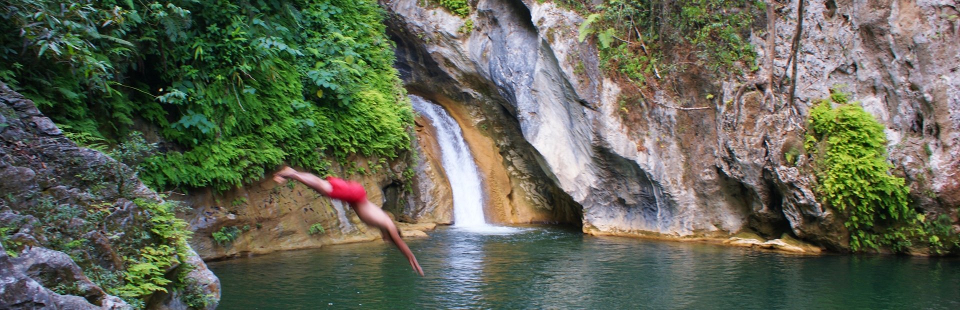 Persoon in rode zwembroek duikt met het hoofd eerst in een natuurlijke poel met waterval en weelderige groene kliffen in Cuba