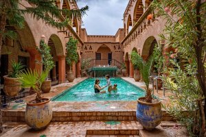Een vrouw en twee kinderen zwemmen in een zwembad op een traditionele binnenplaats met terracotta architectuur en planten in Marokko.