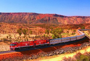 Rood-witte trein van The Ghan die door een droog, rood landschap met lage begroeiing en heuvels rijdt in Australië.