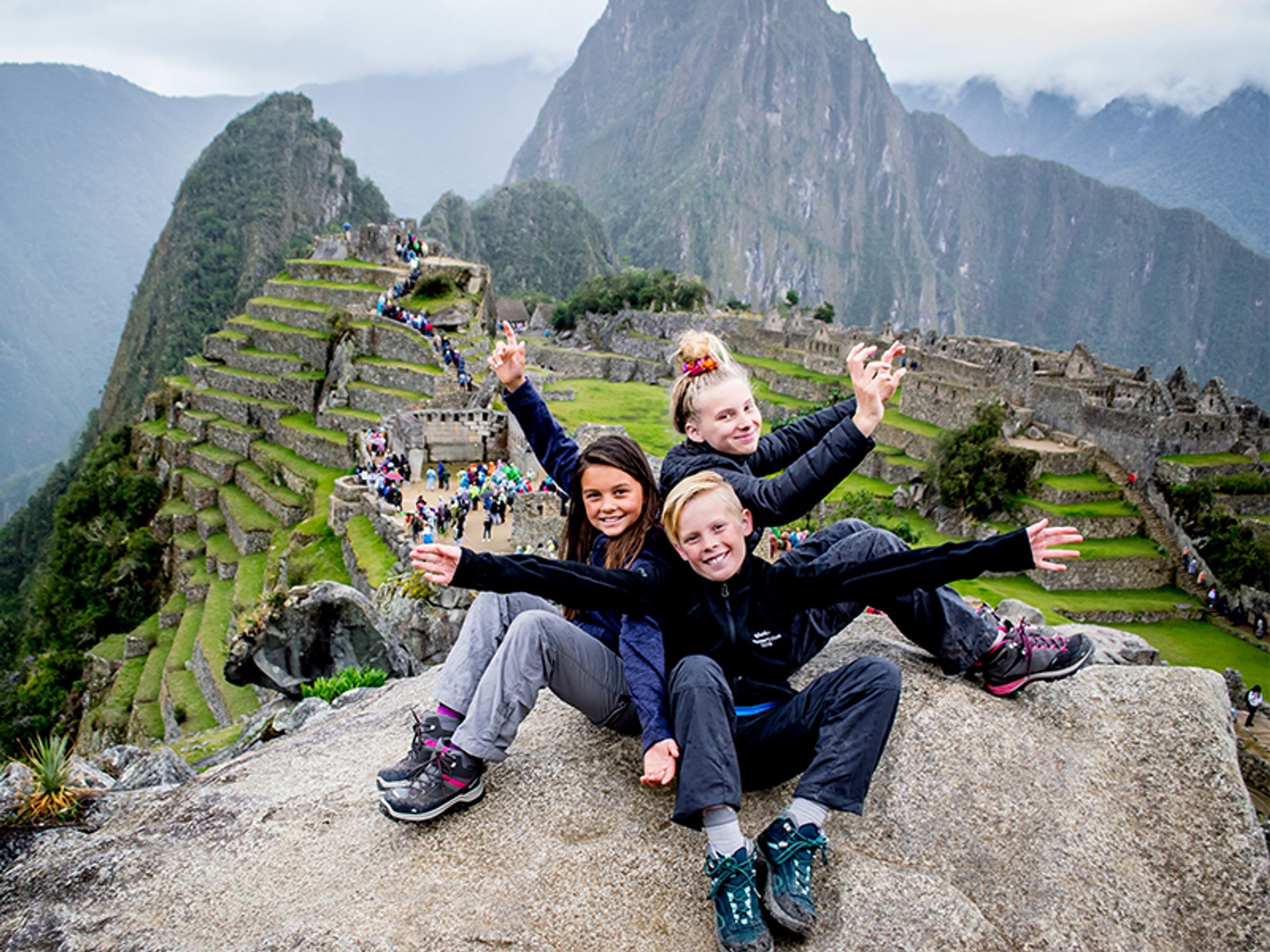 Drie lachende kinderen poseren op een rots met uitzicht op Machu Picchu en de terrasvormige berghelling in Peru.