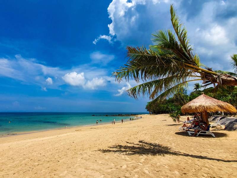 Rustig strand op Koh Lanta met goudgeel zand, helderblauw zeewater en een paar mensen die wandelen of zwemmen. Rechts liggen reizigers ontspannen onder een rieten parasol, omringd door palmbomen die over het strand hangen. De lucht is blauw met wat wolken.