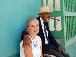 Lachende vrouw met oudere man met hoed die een sigaar rookt en een arm om haar heen slaat tegen een teal muur in Cuba