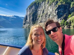 Smiling couple on a boat in a sunny fjord with towering cliffs and a waterfall.