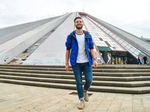 Lachende man in een blauwe jas loopt langs een groot grijs piramidevormig gebouw met glazen panelen en graffiti in Tirana in Albanië