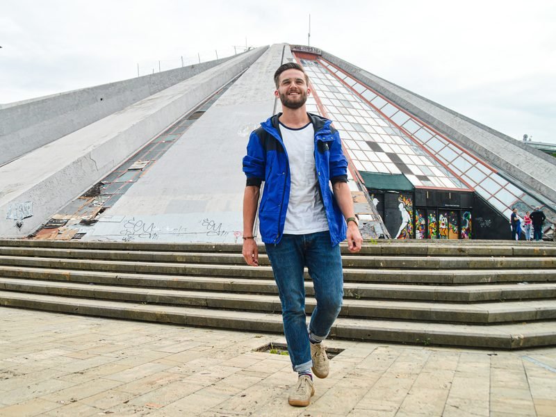 glimlachende man in blauwe jas loopt voor brutalisme Piramide van Tirana Albanië