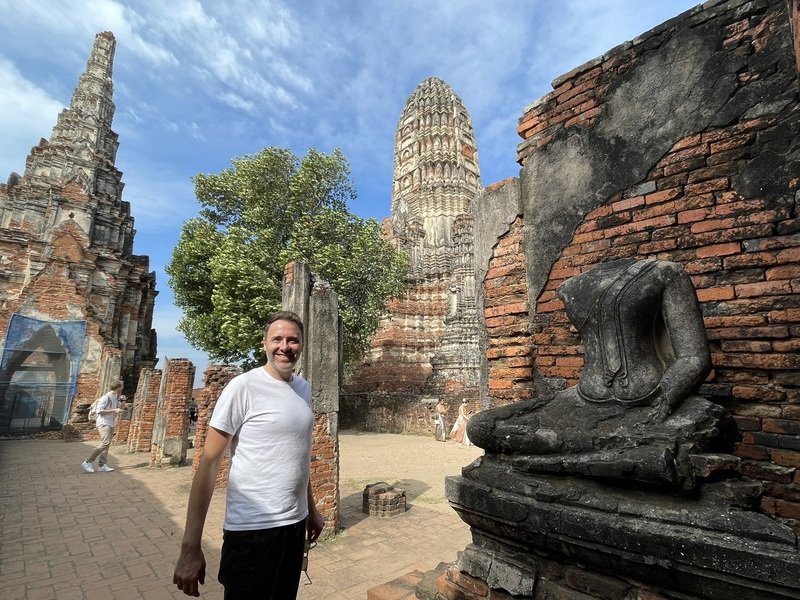 Een man in een wit T-shirt staat lachend tussen eeuwenoude ruïnes van een Boeddhistische tempel in Thailand, Ayutthaya. Op de voorgrond een verweerd, hoofdloos Boeddhabeeld. Op de achtergrond staan indrukwekkende, bakstenen tempeltorens (prangs) met verweerde details. Een grote boom groeit tussen de ruïnes. De lucht is blauw met lichte bewolking.