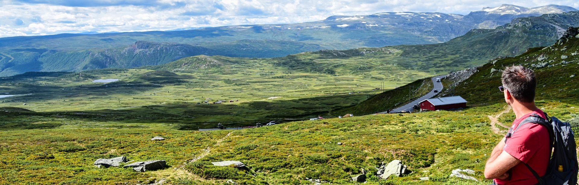A man with a backpack overlooks a vast, green mountainous landscape featuring a winding road, a red building, distant lakes, and snow-capped peaks under a partly cloudy sky.