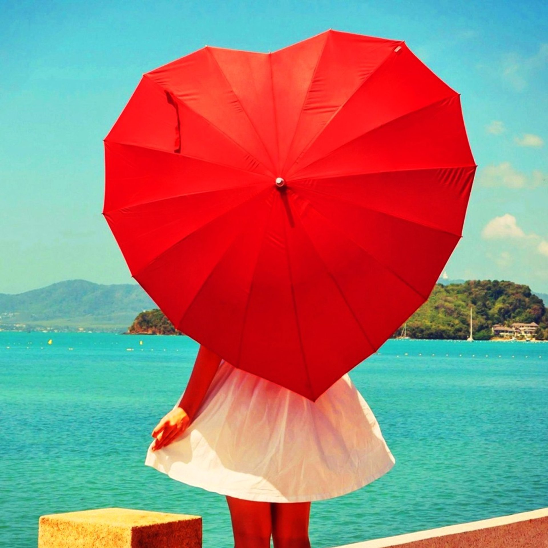 Vrouw in een witte jurk staat aan zee met een groot rood hartvormig paraplu, terwijl op de achtergrond helderblauw water, groene heuvels en een zonnige lucht te zien zijn.