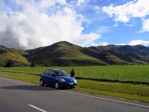 Blauwe auto met een persoon staat langs de weg naast groene velden en glooiende heuvels onder een bewolkte lucht in een landelijk gebied
