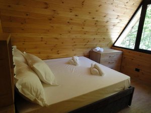 Houten slaapkamer aan het Komanmeer in Albanië met bed onder een schuin dak en een raam met uitzicht op het groen
