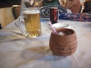 A mug of beer next to a terracotta cup with a straw and ice, on a white tablecloth.