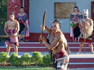 Māori krijger met armtatoeages in traditionele kleding houdt een houten staf vast terwijl anderen in ceremoniële kleding bij een gebouw staan in een culturele setting in Nieuw-Zeeland