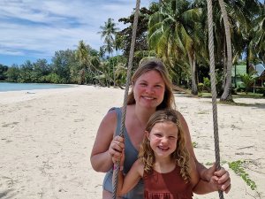 Een lachende vrouw en een meisje zitten op een touwschommel op een zonnig tropisch strand met palmbomen in Thailand.