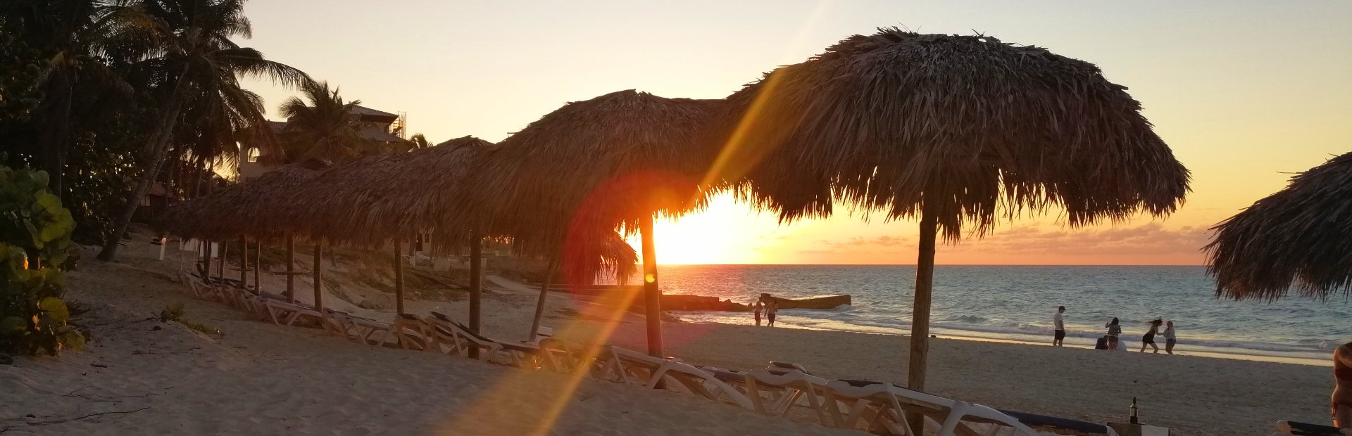 Zonsondergang boven een strand met rieten parasols, ligstoelen en mensen bij de waterlijn in Cuba
