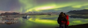 Stel kijkt naar groen noorderlicht dat weerspiegelt in een gletsjerlagune met ijsbergen en bergen in de verte in IJsland