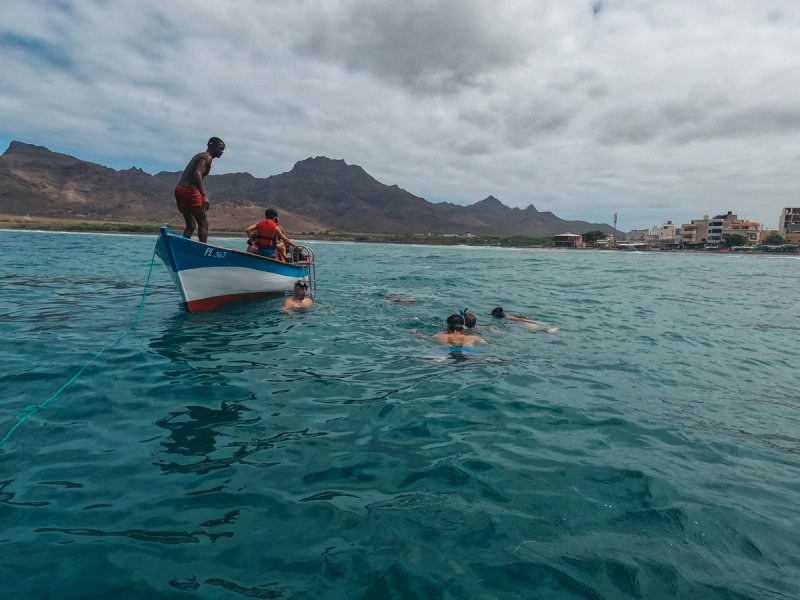 People swim and snorkel around a small boat in blue water with mountains and a town in the distance.