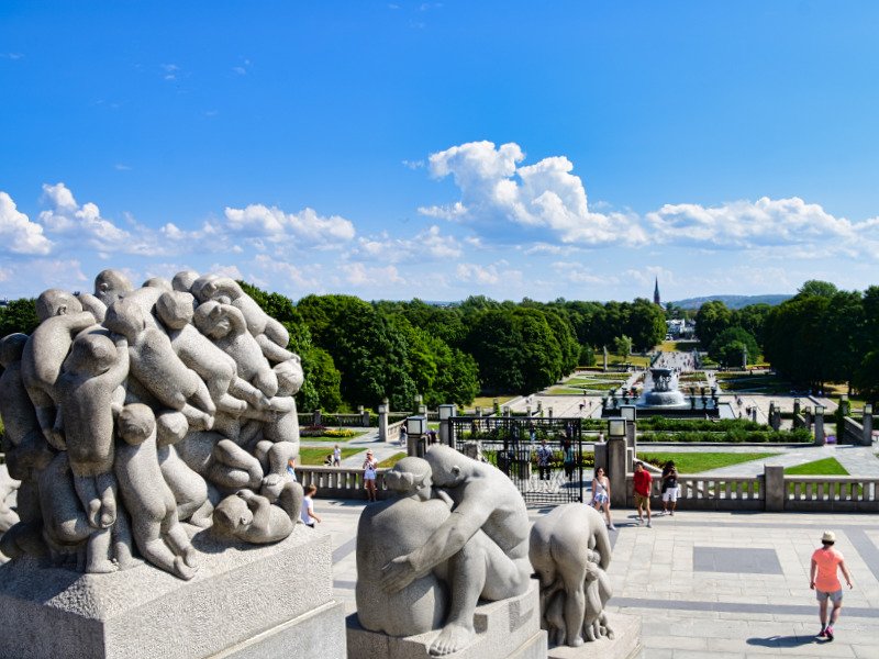 Stone sculptures of intertwined human figures overlook a vast, green park with fountains and trees under a blue sky.