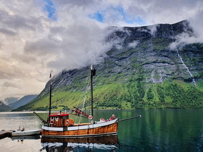 A wooden boat on calm water, with a green mountain partially obscured by clouds and waterfalls.