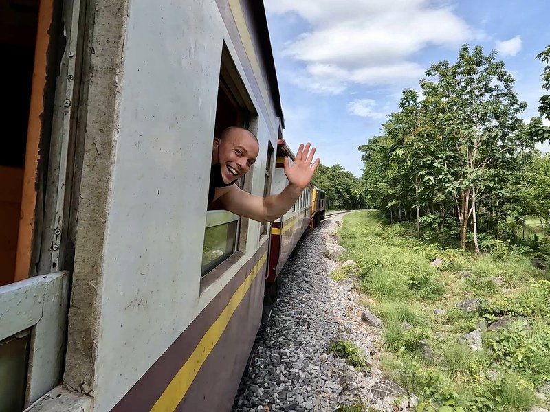 Reiziger steekt lachend zijn hoofd en hand uit het raam van een rijdende trein die door een groen landschap rijdt, met bomen en gras langs het spoor.