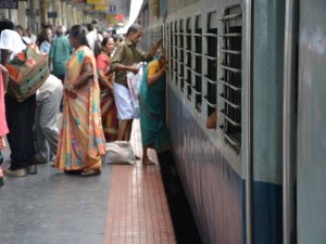 Mensen in kleurrijke kleding op een druk treinstation in India, waar een vrouw door het raam van een blauwe trein naar binnen reikt.