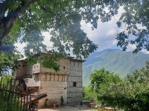A traditional stone building with wooden elements, framed by green leaves and trees, against a backdrop of lush green mountains under a cloudy sky.