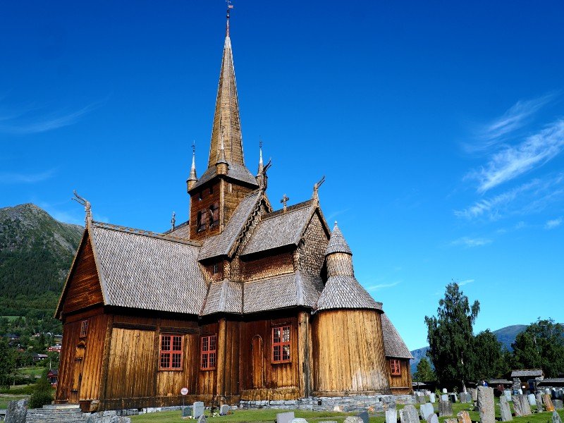 A historic brown wooden stave church with a tall spire stands next to a graveyard under a bright blue sky.