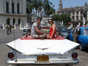 Lachend stel zit op de kofferbak van een witte klassieke auto uit de jaren 50 met opvallende staartvinnen op een zonnige straat met historische gebouwen op de achtergrond in Cuba