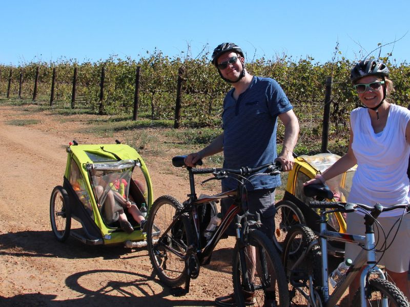 Koppel met helmen staat met fietsen en kindertrailers, waarvan één met een kind erin, op een zandpad langs een wijngaard in Zuid-Afrika