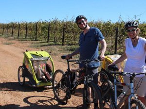 Koppel met helmen staat met fietsen en kindertrailers, waarvan één met een kind erin, op een zandpad langs een wijngaard in Zuid-Afrika