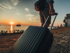 Reiziger met koffer loopt over een stenen pad langs de kust bij zonsondergang, met een schip zichtbaar op zee.