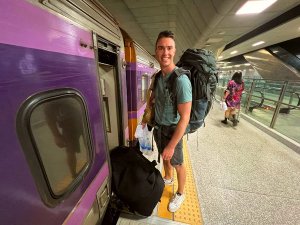 Reiziger met een grote backpack bij de ingang van een paarse trein op een fel verlicht treinstation.