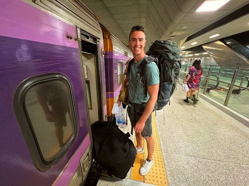 Reiziger met een grote backpack bij de ingang van een paarse trein op een fel verlicht treinstation.