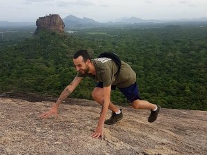 Man klimt een rots op met uitzicht over een uitgestrekt groen boslandschap en de rotsformatie Sigiriya in Sri Lanka op de achtergrond.