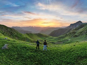Twee mensen op een groene berghelling in Albanië kijken naar de zonsondergang over een uitgestrekt berglandschap