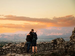 Twee wandelaars kijken bij zonsondergang uit over een bergketen en een stad in Albanië, terwijl de man wijst