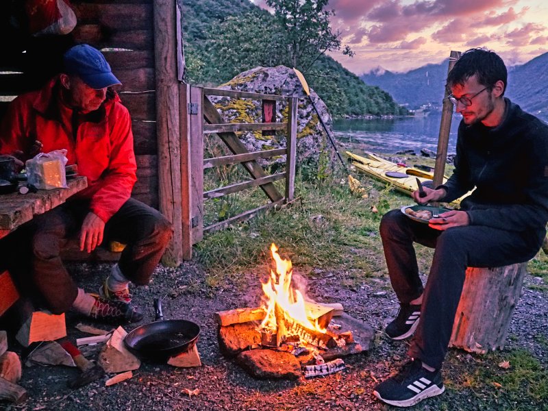 Two men sit by a campfire near a fjord with a cabin and kayaks, under a sunset sky.