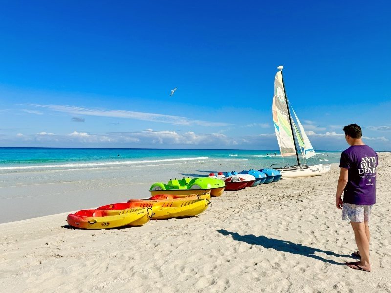 Een man op een wit zandstrand met kleurrijke kajaks, waterfietsen en een zeilboot, gericht naar een turquoise oceaan onder een helderblauwe lucht in Cuba